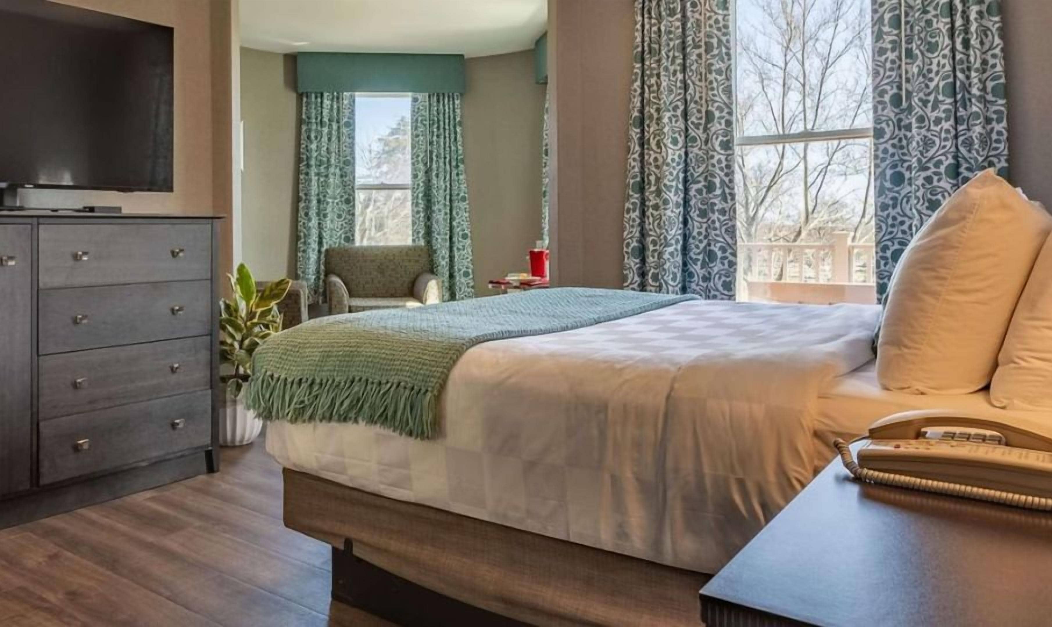 Een slaapkamer met een bed, een dressoir, een stoel, een televisie en een raam met gordijnen.
