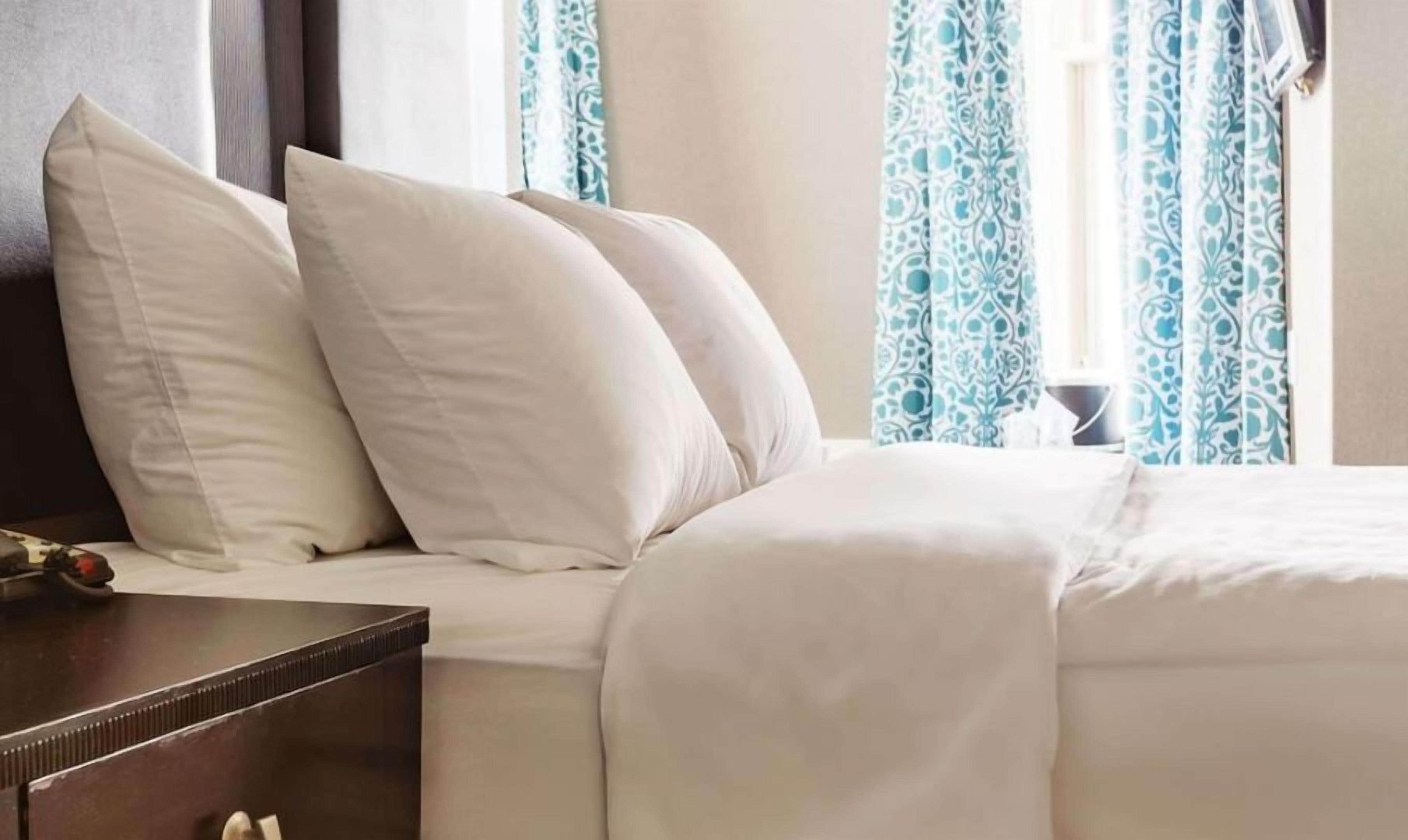 Una cama bien hecha con ropa de cama y almohadas blancas, una mesita de noche de madera oscura con un control remoto y una ventana con cortinas de estampado azul.