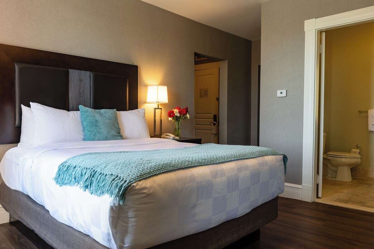 Una cama bien hecha con ropa de cama blanca y verde azulado, una lámpara de noche y un florero con flores rojas.