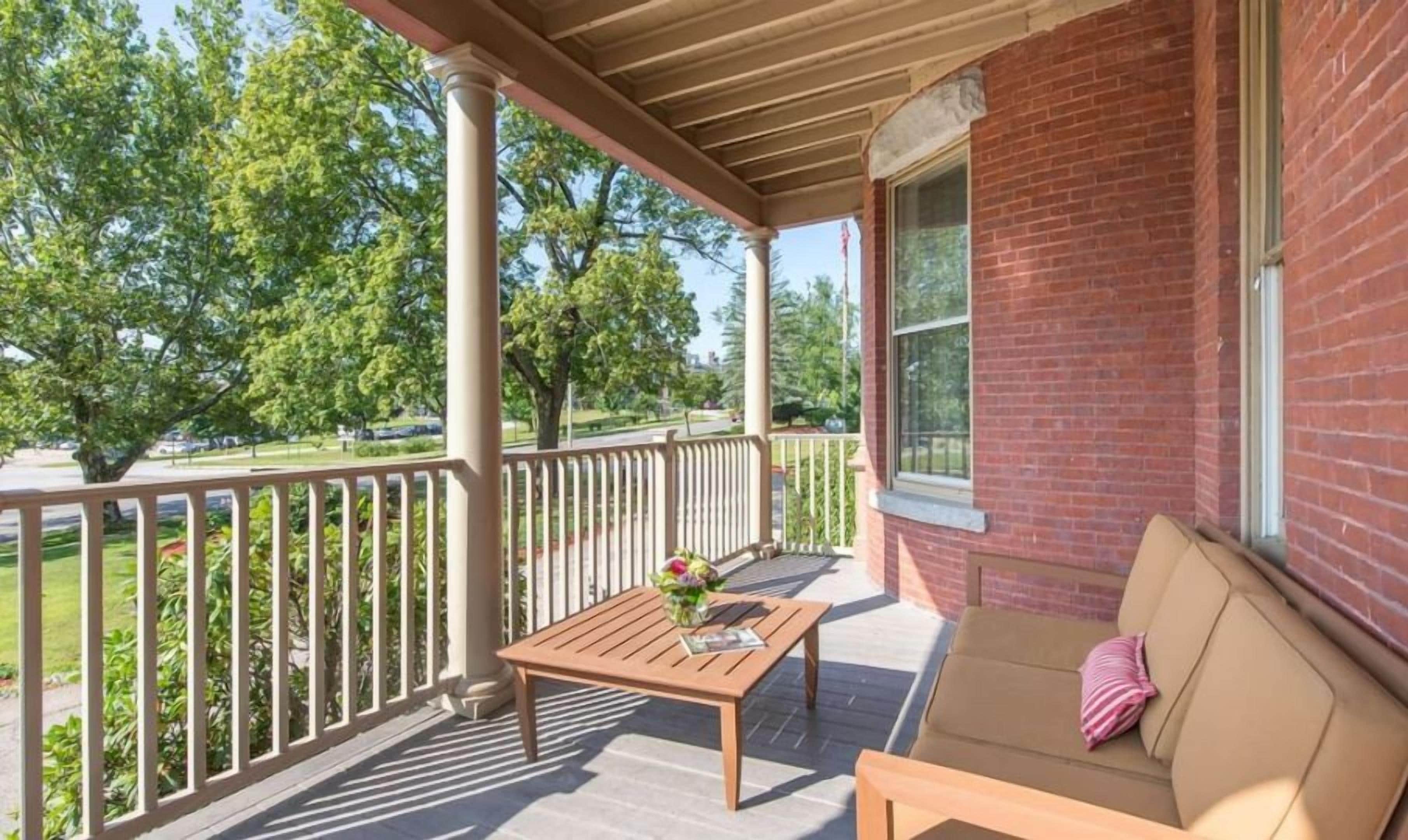 A covered porch with a wooden table and a sofa, a brick wall, and a view of trees and a road.