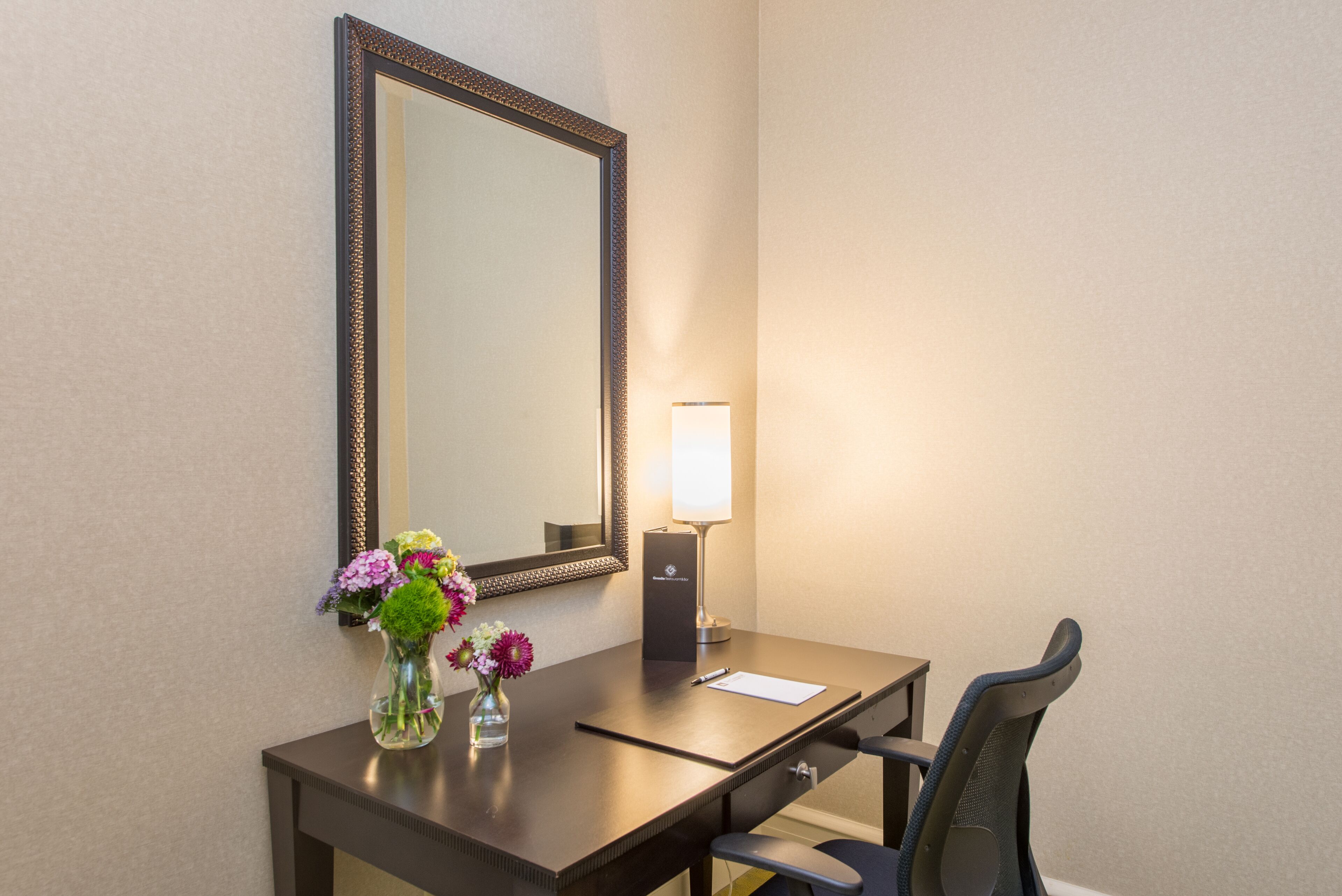 A hotel room with a desk, chair, mirror, vase with flowers, and a lamp.