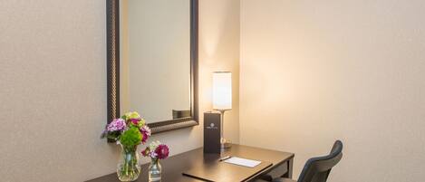 A hotel room with a desk, chair, mirror, vase with flowers, and a lamp.