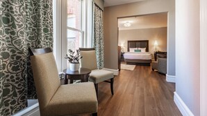 A hotel room with a bed, bedside tables, a chair, a small table with a plant, and a window with patterned curtains. - The Centennial (Concord)