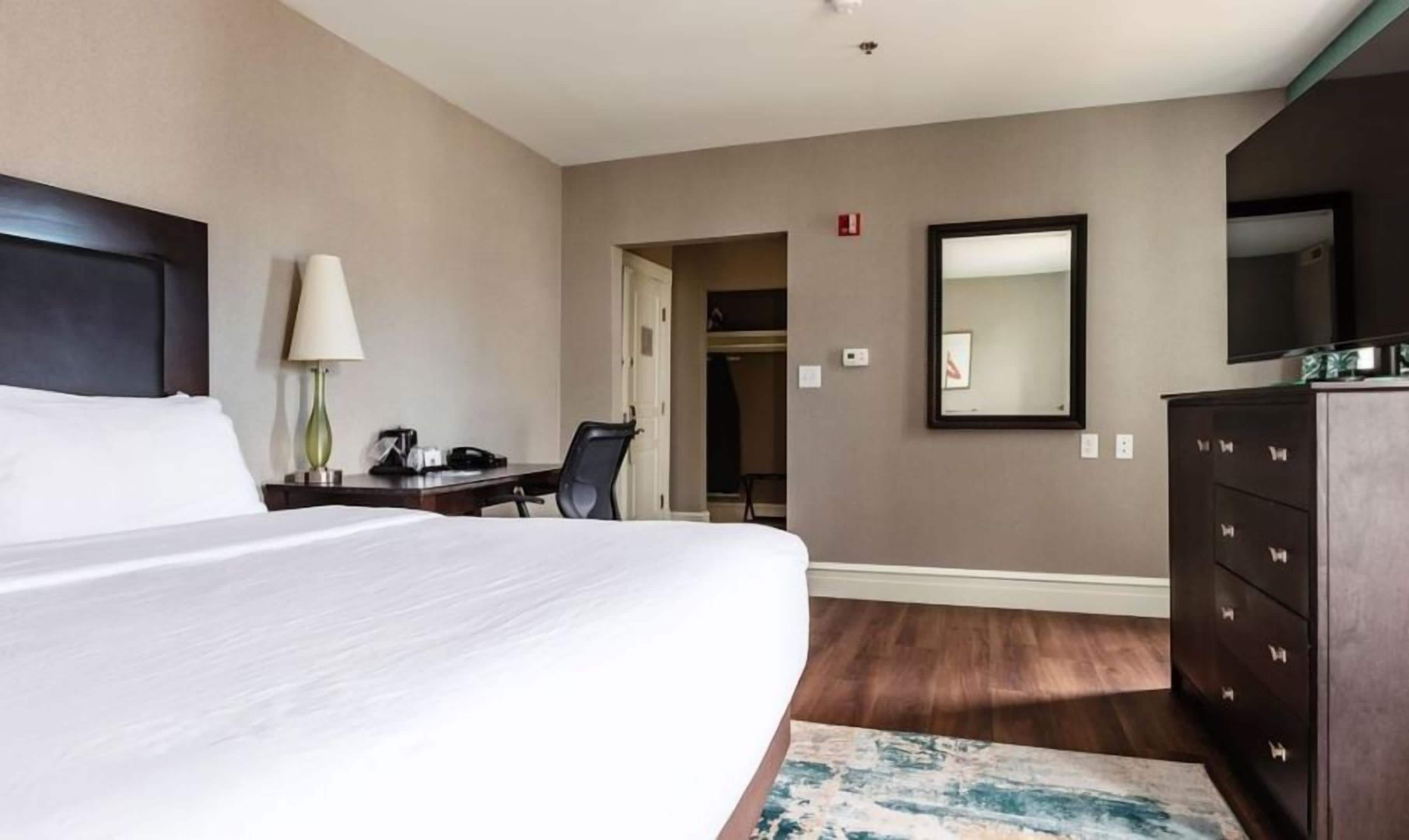 A hotel room with a large bed, a desk with a lamp, a dresser, and a mirror on the wall.