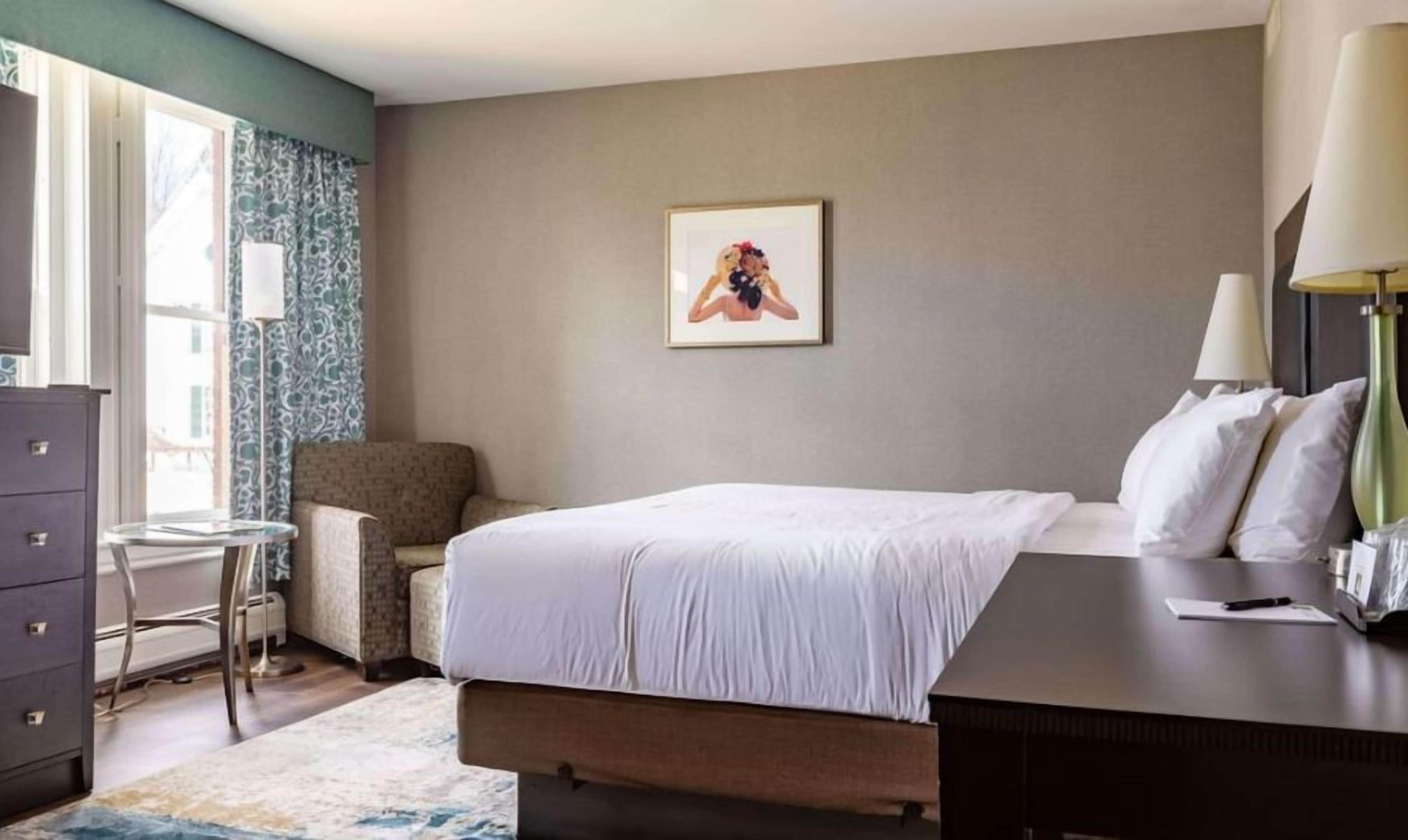 Een hotelkamer met een bed, een bureau, een stoel, een dressoir, een lamp en een lijst aan de muur.