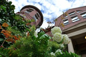 A building with a brick facade and a circular window, surrounded by lush greenery and white flowers.