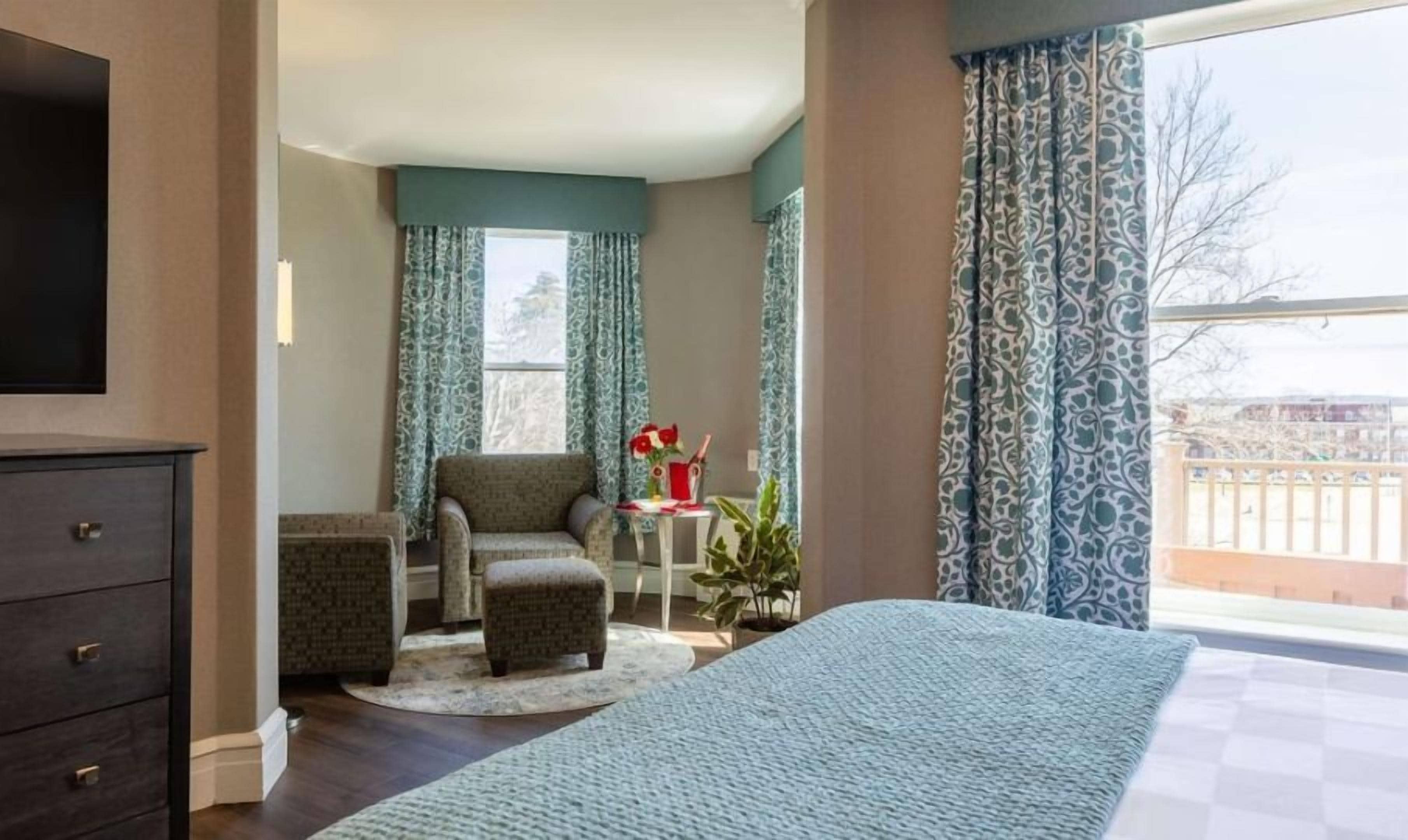 A hotel room with a bed, a chair, a small table with a vase, and a view of the outside.