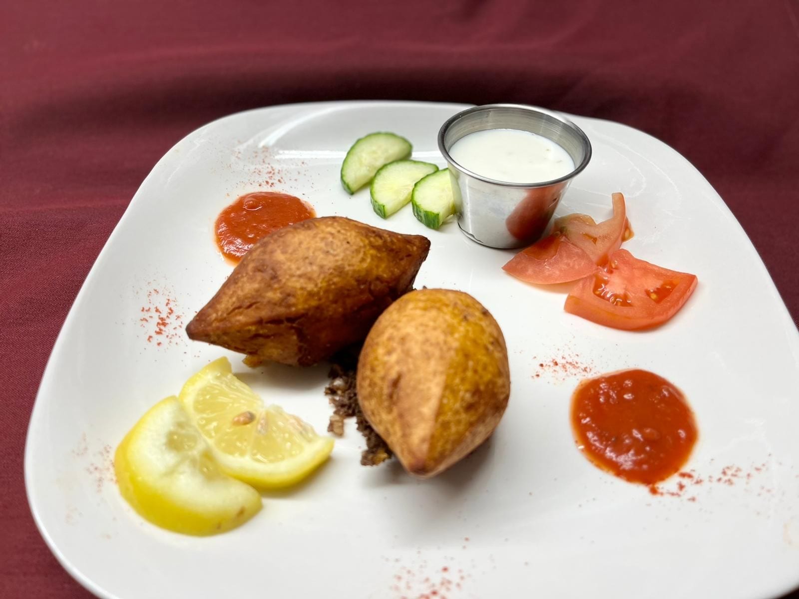 A plate with samosas, cucumber slices, tomato slices, lemon wedges, and two types of sauce.