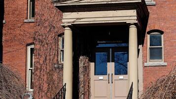 A red brick building with a beige entrance, a set of stairs leading up to the door, and a black metal railing on the left.