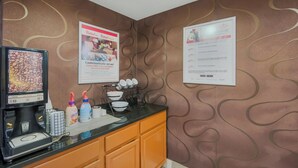 Una estación de café con una máquina de café, tazas y botellas de condimentos. Hay dos carteles en la pared.