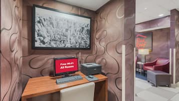 A hotel room with a desk, a computer, and a printer. There is a framed cityscape picture on the wall.