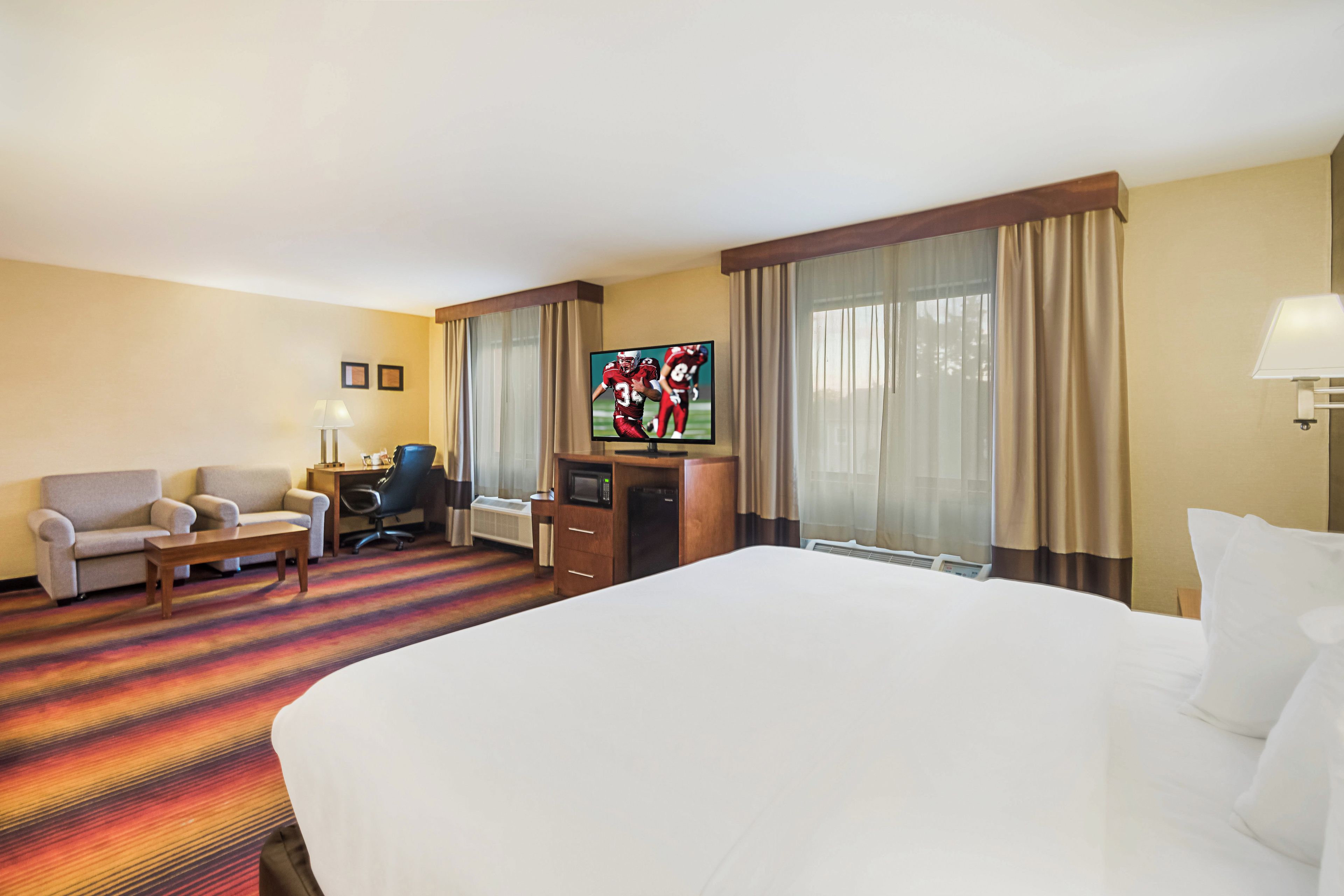 A hotel room with a large bed, a desk with a computer, a chair, a TV, and a window with curtains.