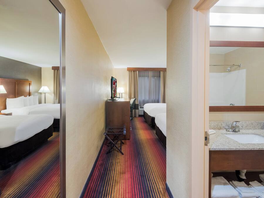 Habitación de hotel con una cama grande, un escritorio con una silla, un baño con lavamanos y espejo, y una alfombra colorida a rayas.