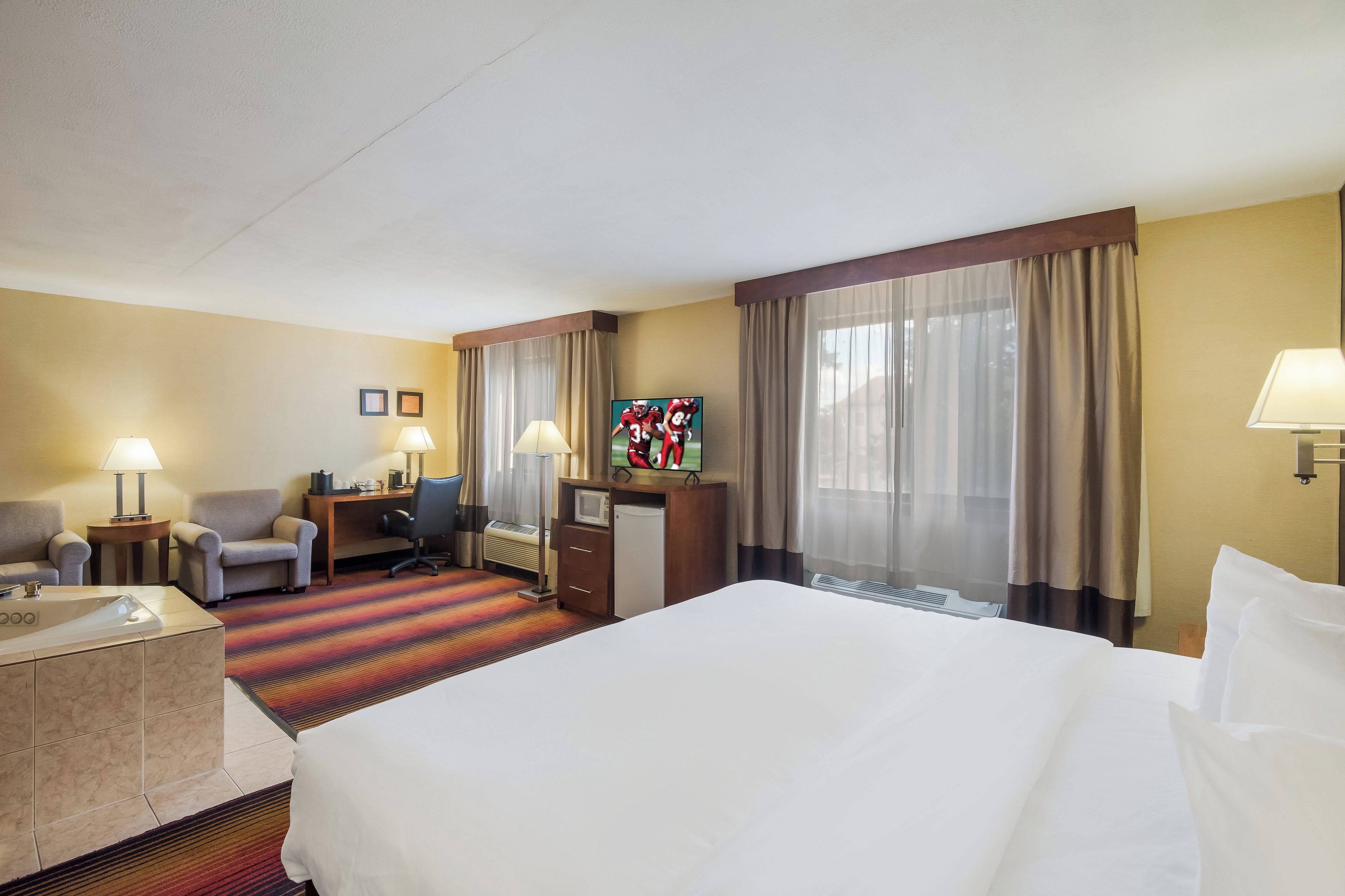 Una habitación de hotel con una cama grande, dos sillones, un escritorio con lámpara, televisor y ventana con cortinas.