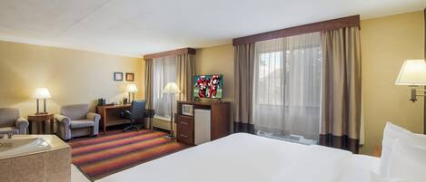 Una habitación de hotel con una cama grande, dos sillones, un escritorio con lámpara, televisor y ventana con cortinas.