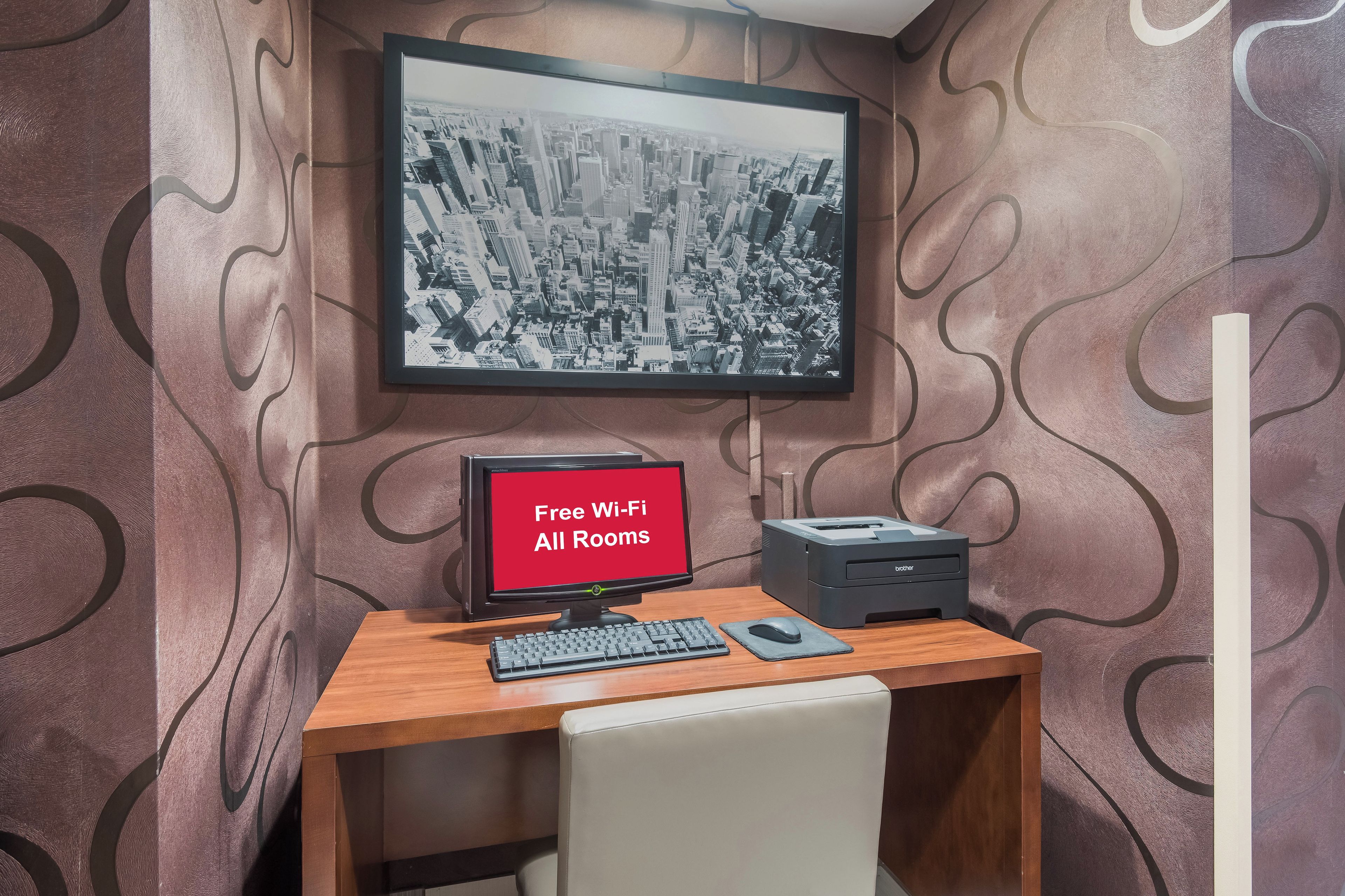 A room with a desk, a computer monitor, a printer, and a framed cityscape picture on the wall.