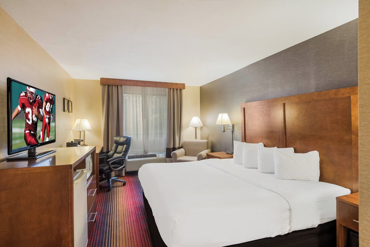 A hotel room with a large bed, a desk with a TV, two chairs, a desk lamp, a nightstand, and a window with curtains.