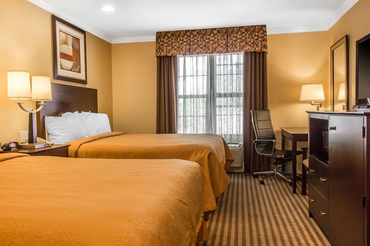 A hotel room with two beds, a desk, a chair, a TV, and a large window with curtains.