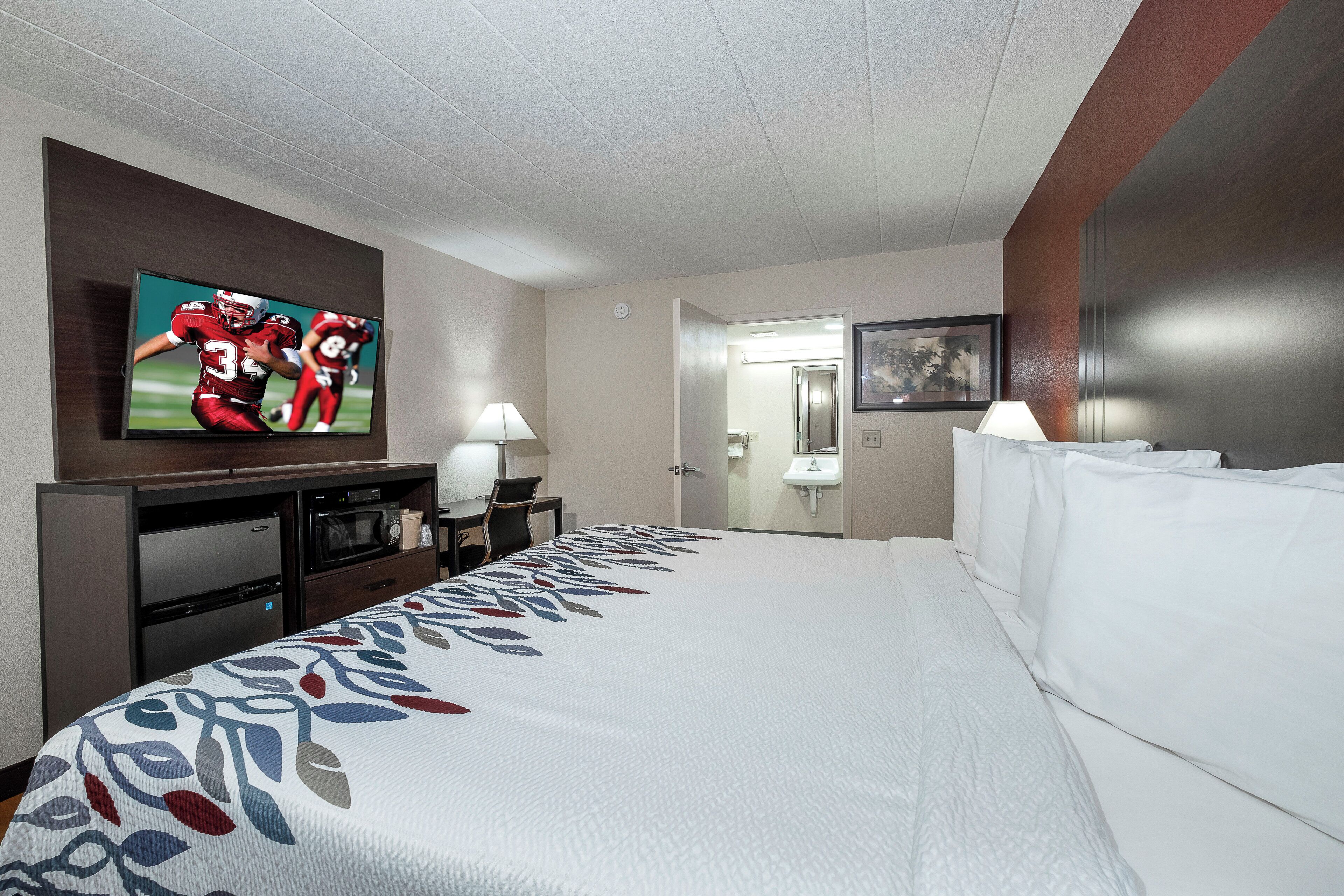 Habitación de hotel con una cama grande, un televisor de pantalla plana en la pared, un escritorio con lámpara, microondas y refrigerador.
