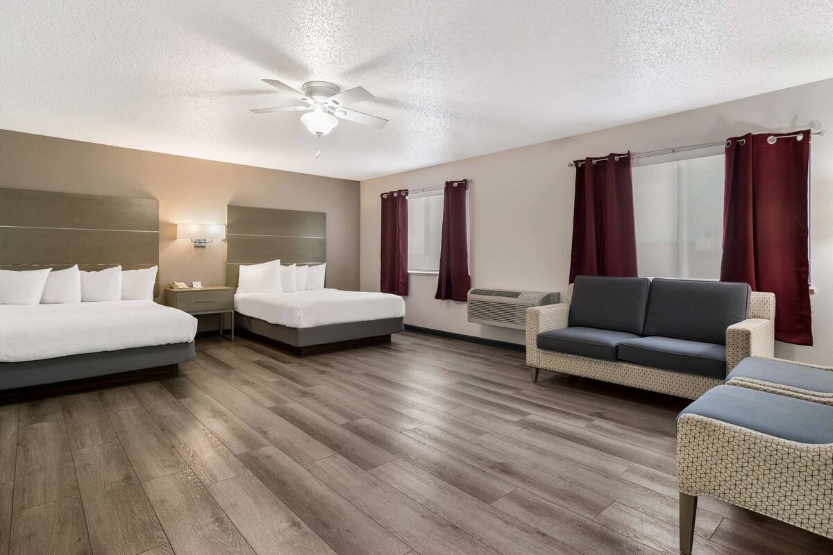 A hotel room with two beds, a sofa, a chair, a ceiling fan, and a window with curtains.