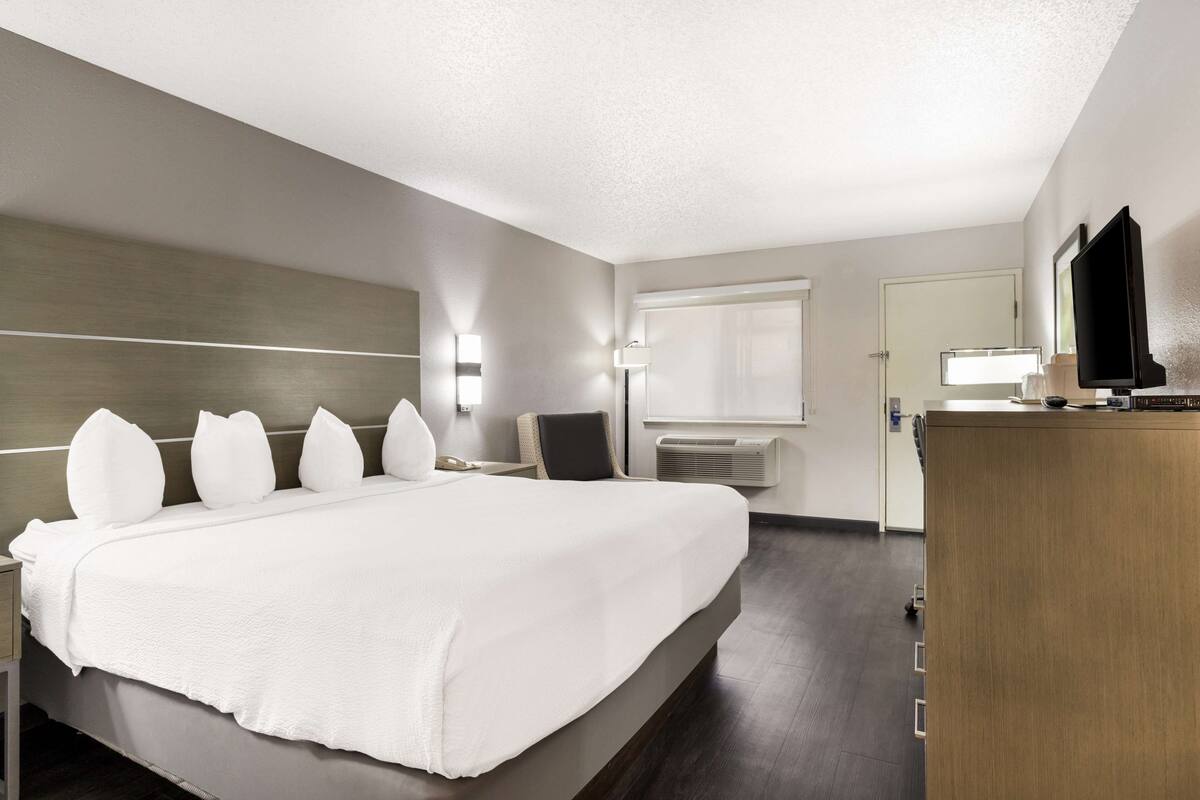A hotel room with a large bed, bedside tables, a desk, a chair, a flat-screen TV, and a window with blinds.