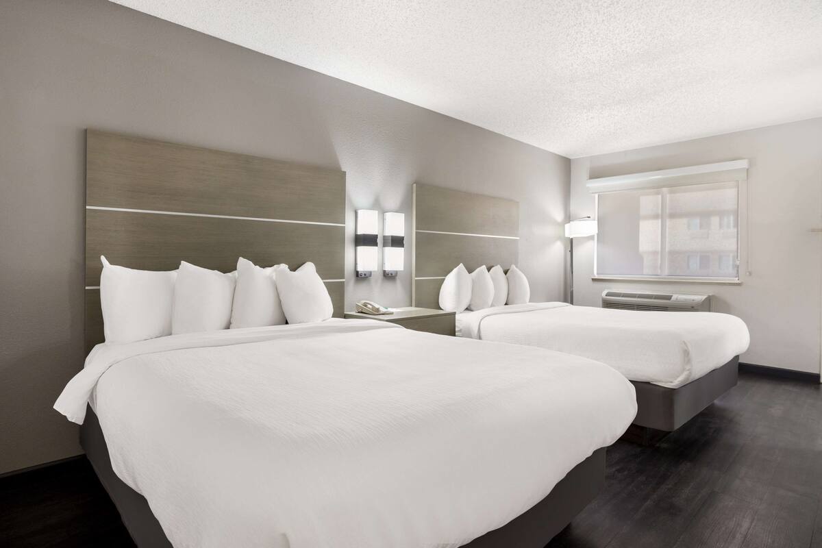A hotel room with two beds, a large mirror, a bedside table with a lamp, and a window with blinds.