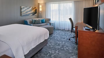 A hotel room with a large bed, a sofa, a desk with a TV, and a window with curtains.
