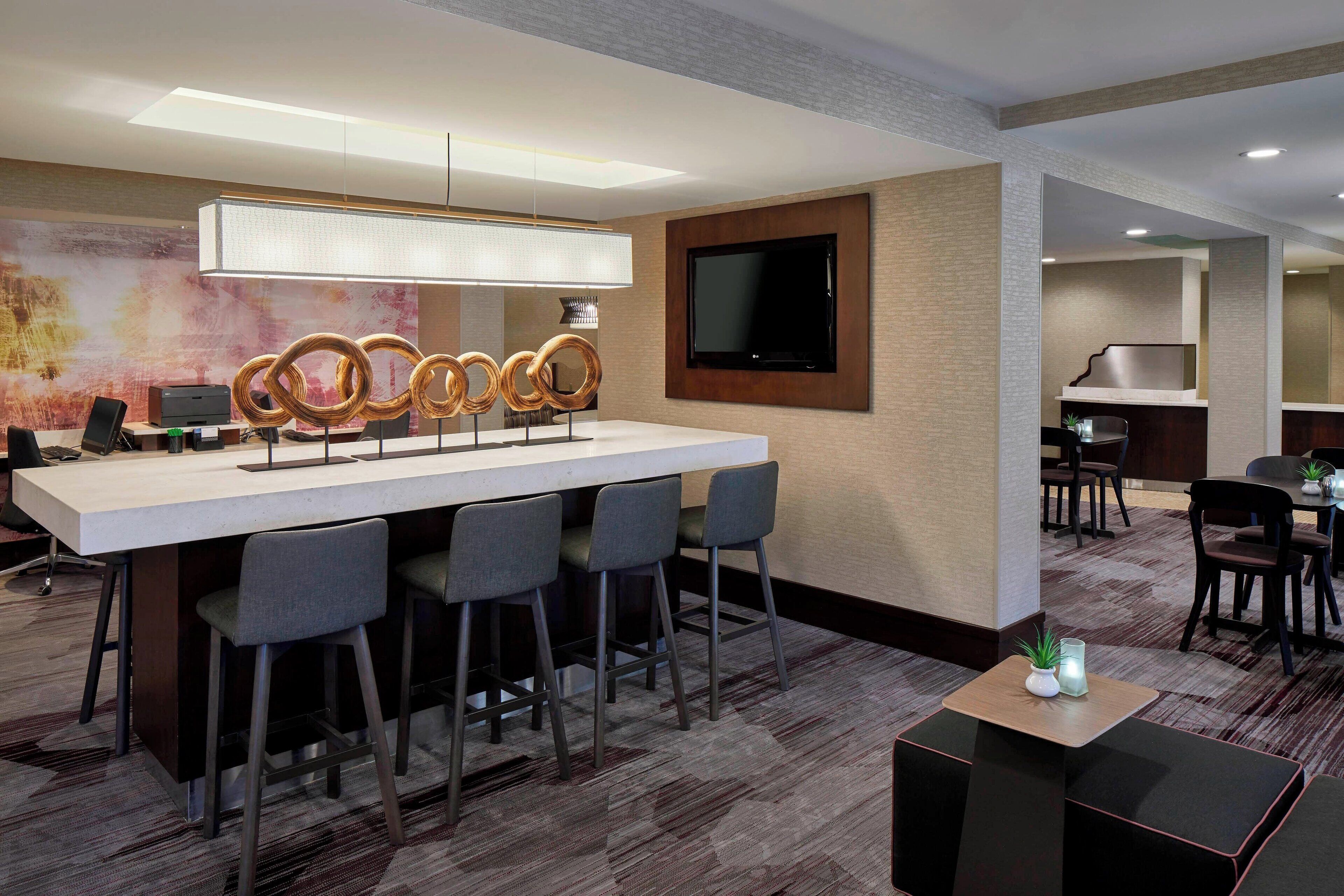 Un bar de hotel moderno con una barra alargada, varios taburetes, un televisor de pantalla plana y una pared decorativa con arte abstracto.