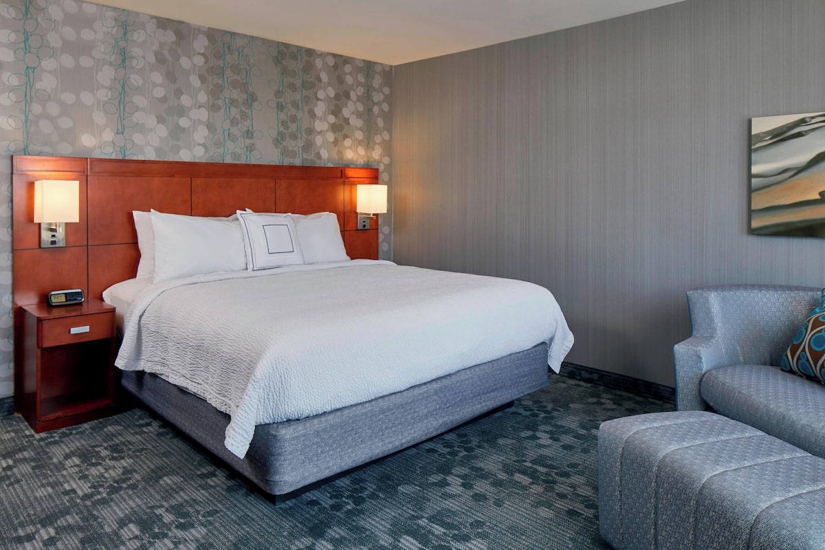 Habitación de hotel con una cama grande, dos mesitas de noche con lámparas, un sillón gris y una alfombra estampada.