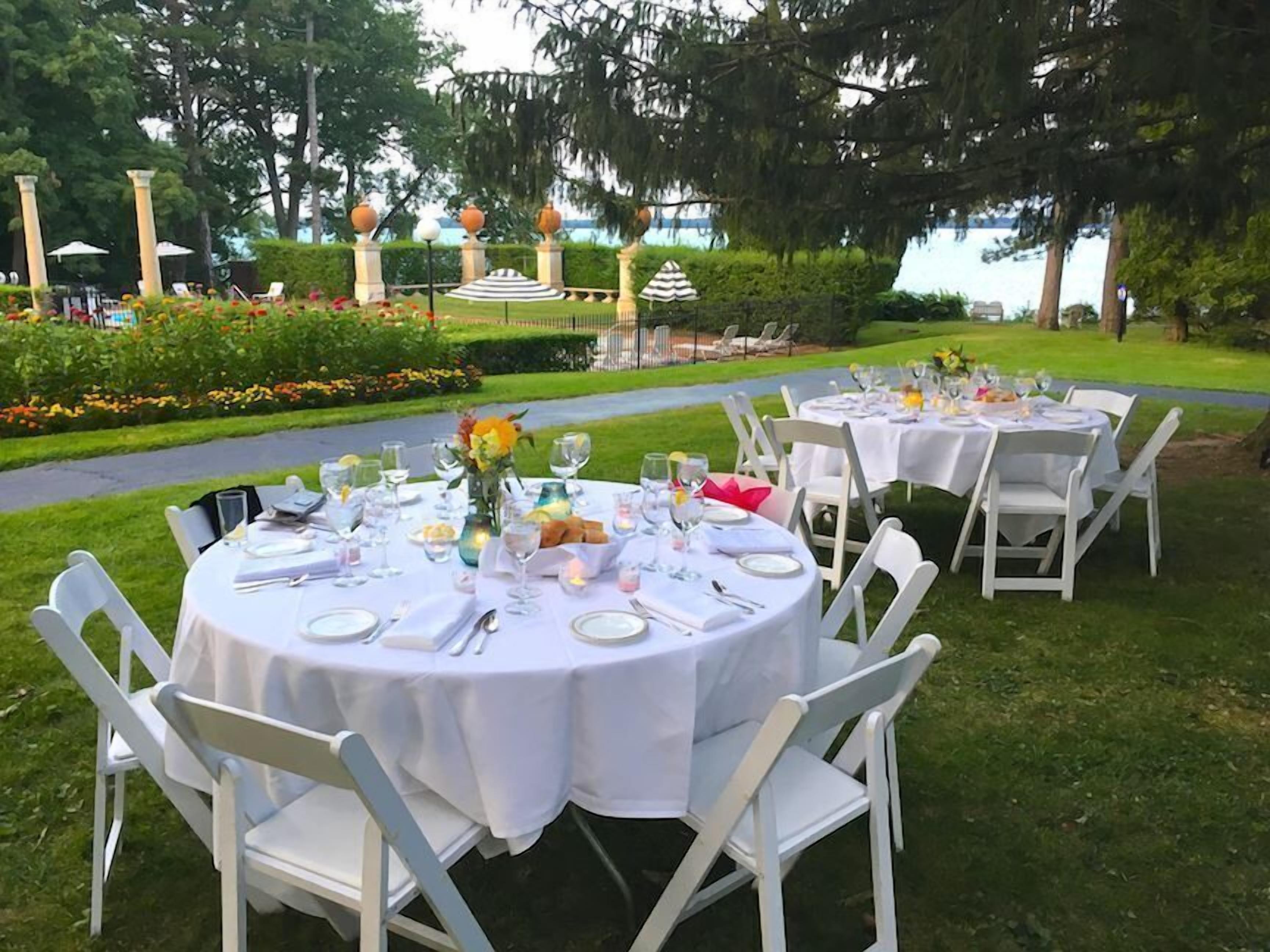 Outdoor round tables set up with white tablecloths and chairs, arranged in a garden setting with a view of a body of water and trees in the background.