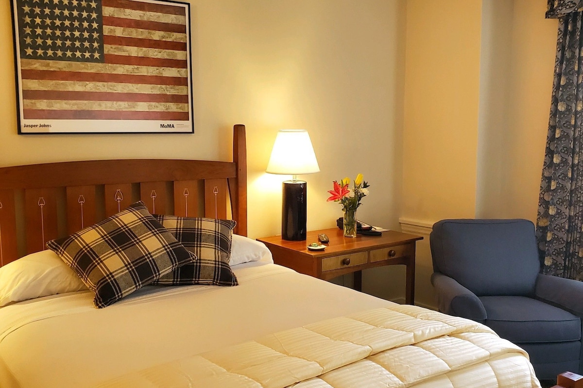 A hotel room with a bed, a nightstand with a lamp, a chair, a wall-mounted American flag, and a window with curtains.