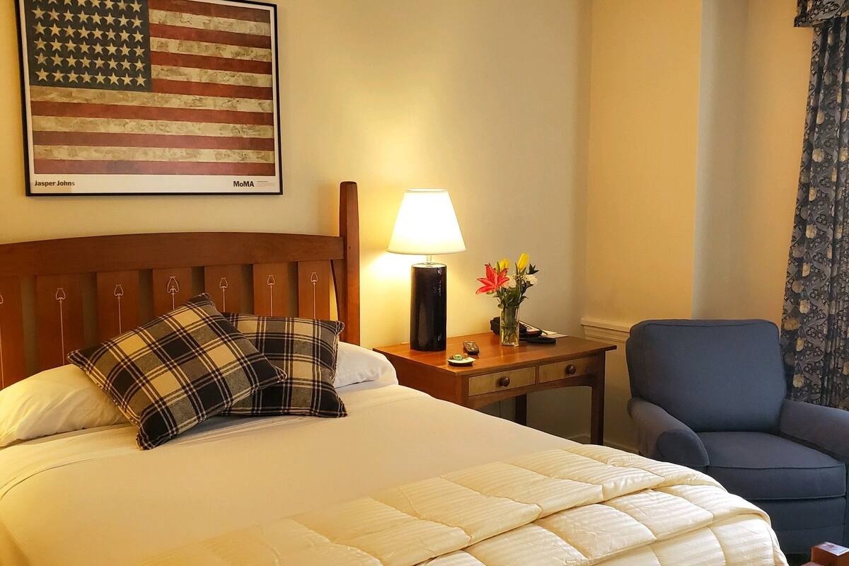 A hotel room with a bed, a nightstand with a lamp, a chair, a wall with an American flag, and a window with curtains.