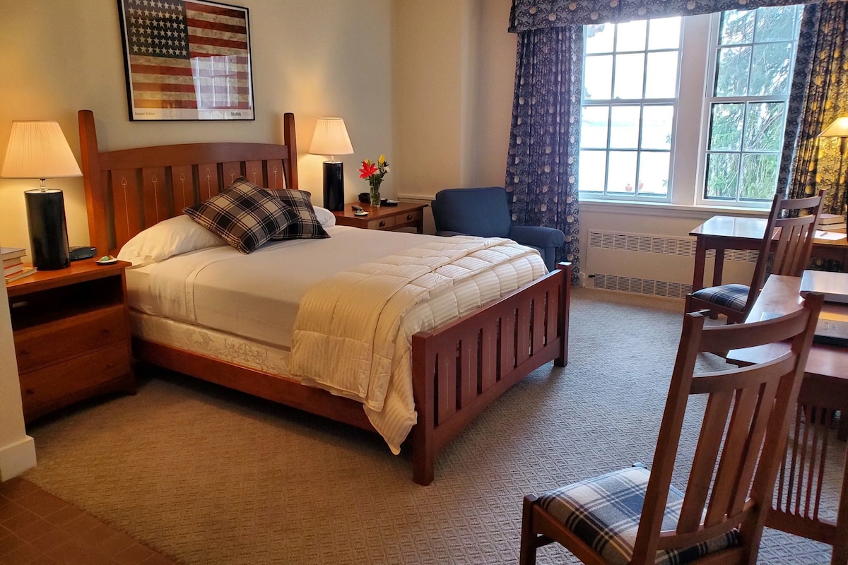 A hotel room with a bed, two bedside lamps, a chair, a table, a window with curtains, and an American flag on the wall.