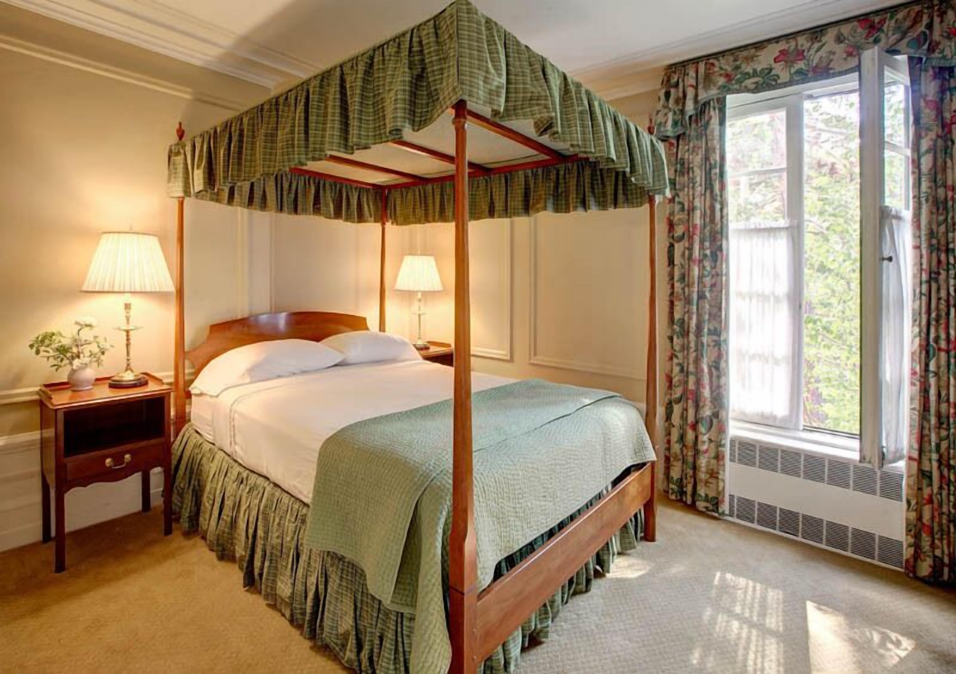 Una cama con dosel sobre cuatro postes, con una colcha y cortinas a juego en color verde, dos mesitas de noche con lámparas y una ventana con cortinas florales.