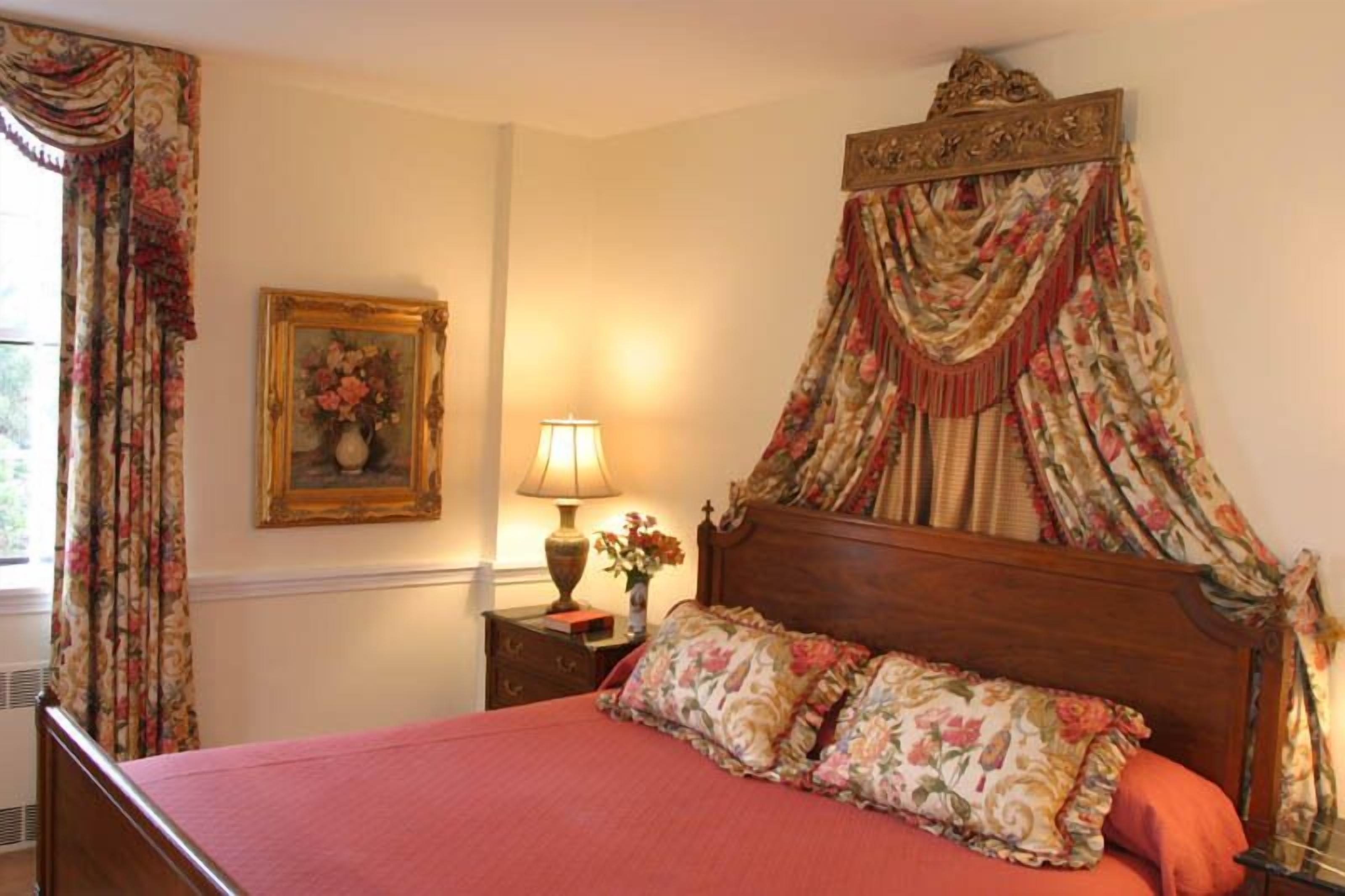 A bedroom with a canopy bed, floral curtains, a bedside lamp, and a framed floral painting on the wall.