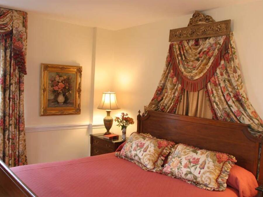 Ein Schlafzimmer mit Himmelsbett, blumigen Vorhängen, einer Nachttischlampe und einem gerahmten Blumenbild an der Wand.