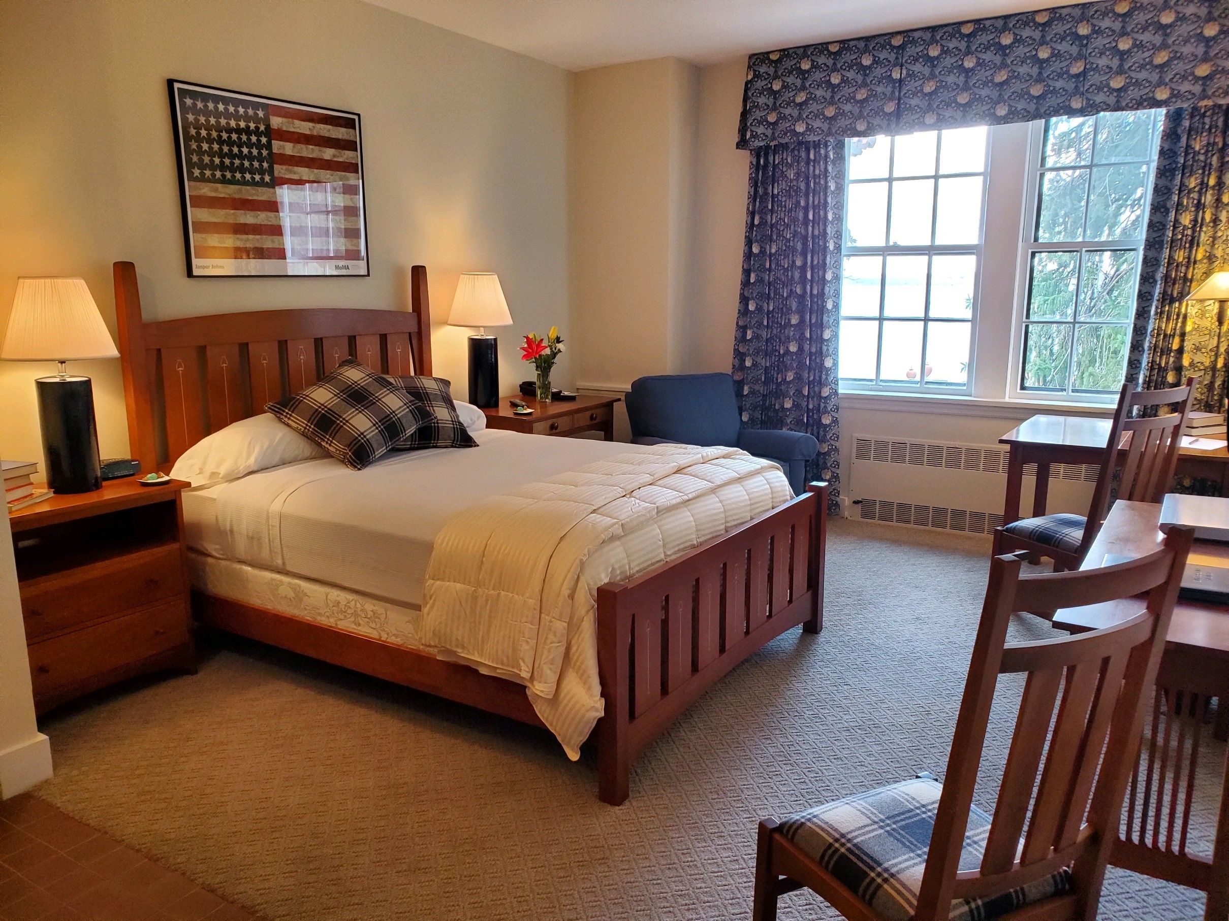 A neatly arranged bedroom with a bed, bedside lamps, a chair, a table, and a window with curtains.