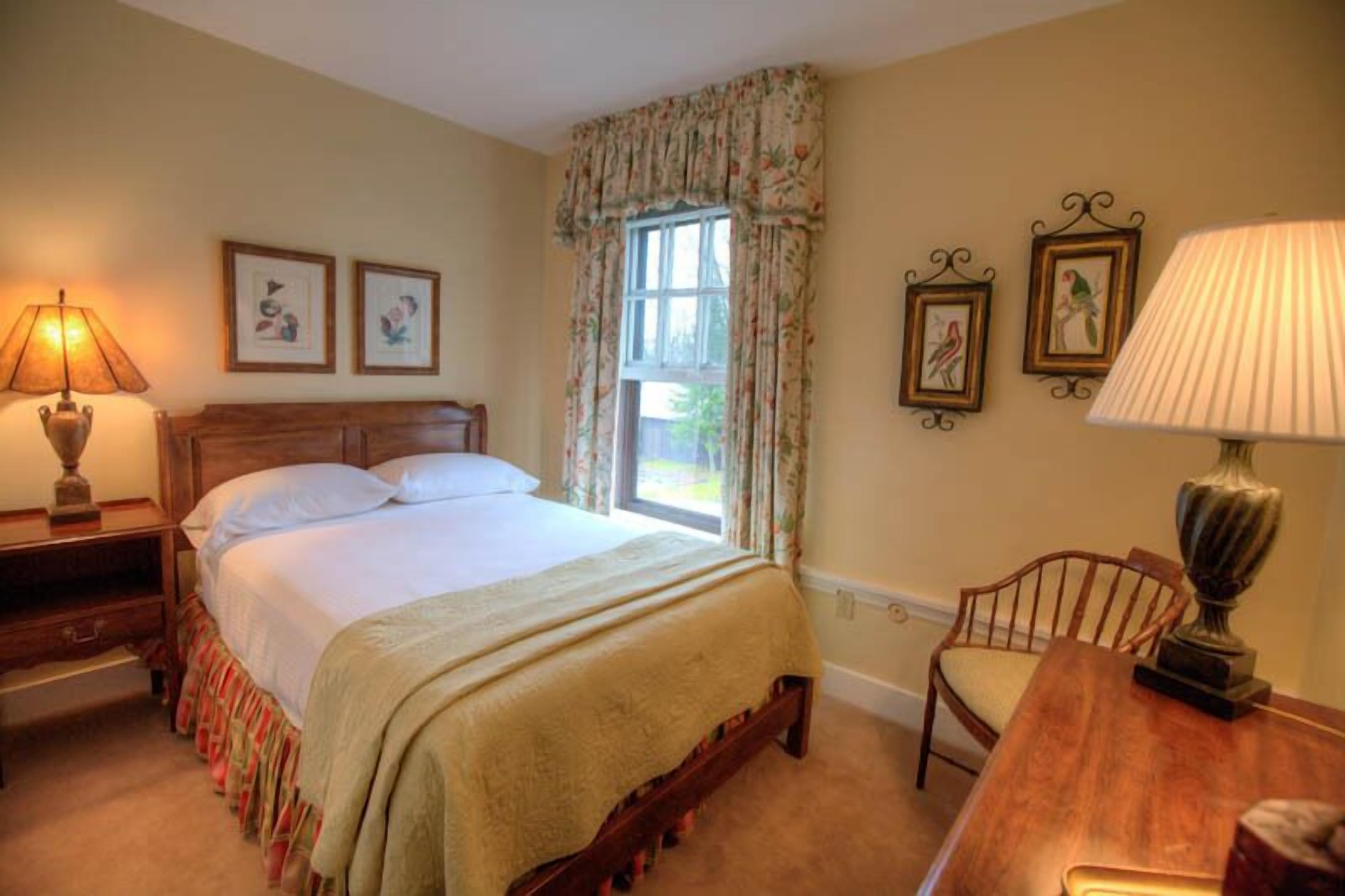 A bedroom with a bed, a nightstand, a chair, a table, and a window with curtains.