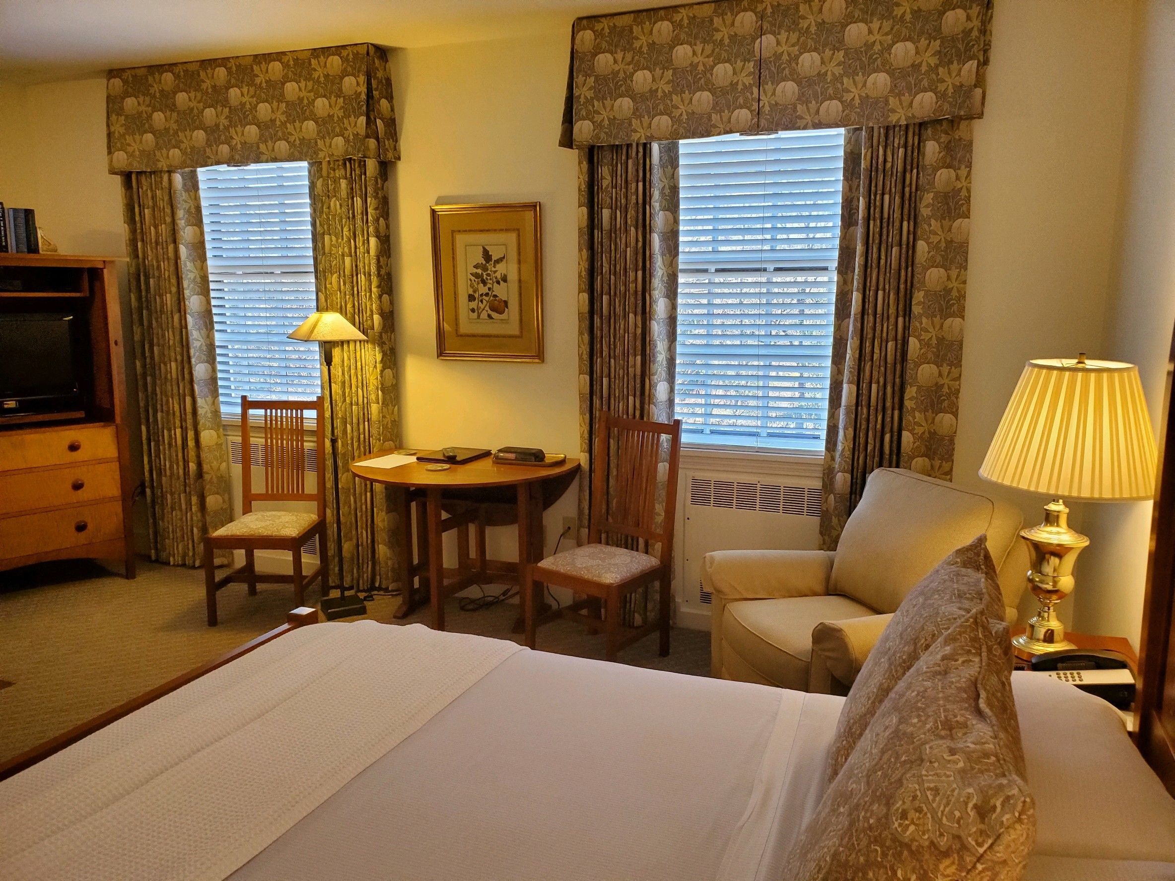 A hotel room with a bed, two chairs, a small table, a desk, a television, and a lamp.