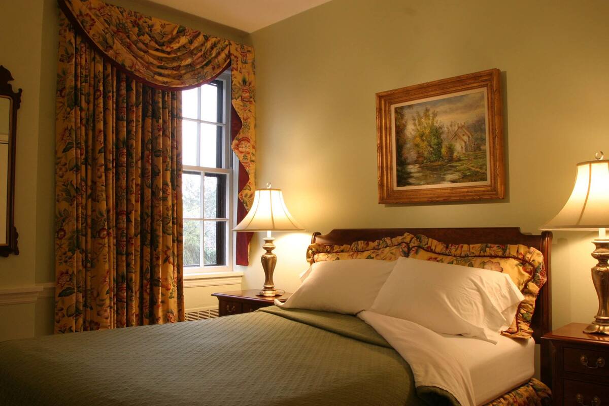 A neatly made bed with floral bedding, a painting on the wall, and a window with curtains.