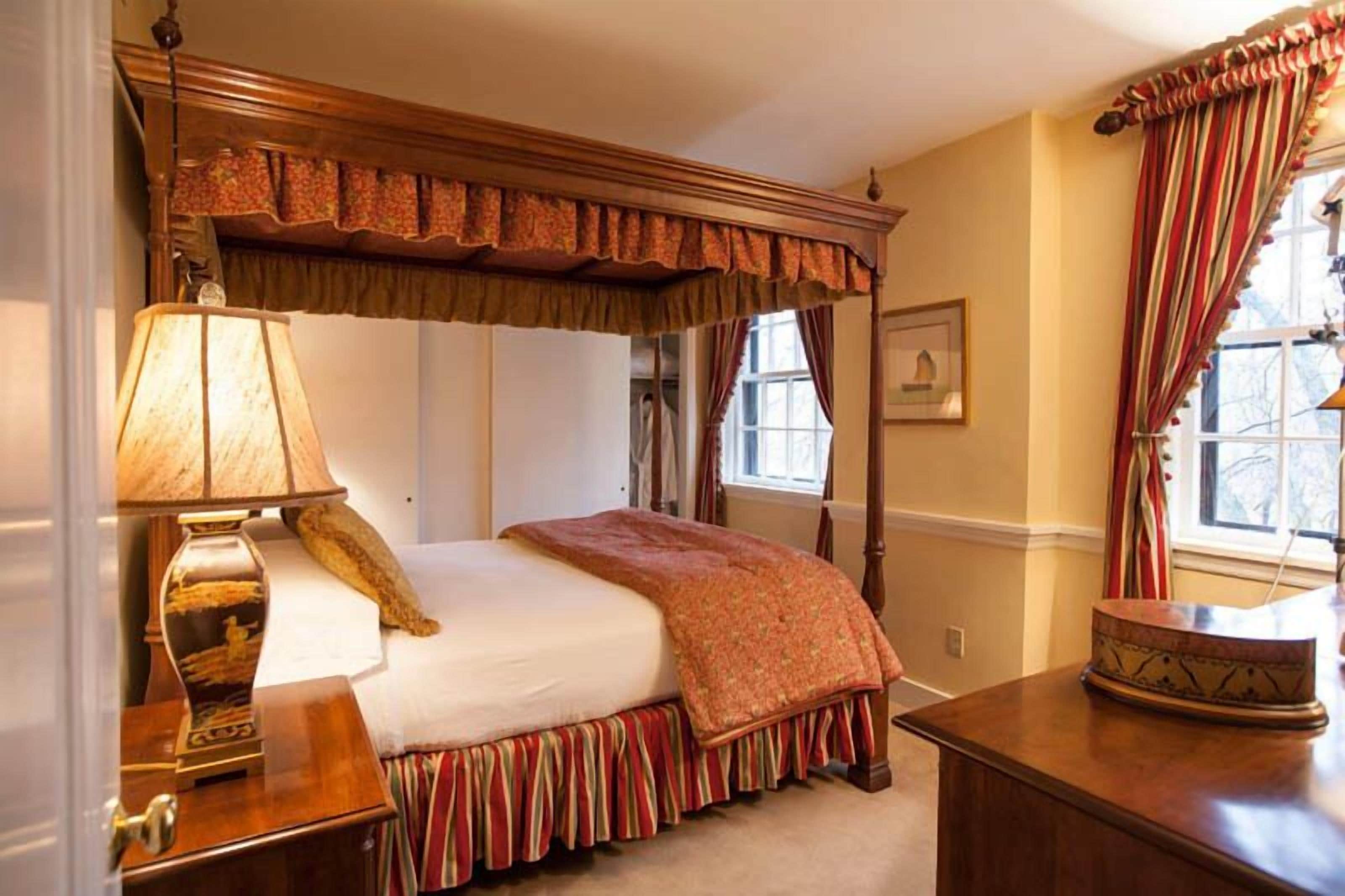A traditional bedroom with a canopy bed, a bedside table with a lamp, and a window with curtains.