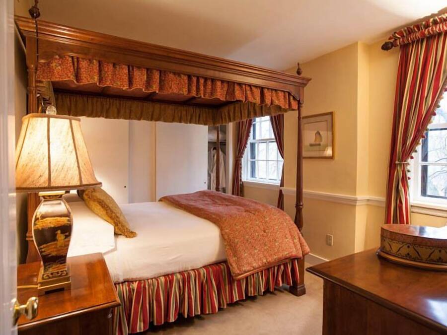 A traditional bedroom with a canopy bed, a bedside table with a lamp, and a window with curtains.