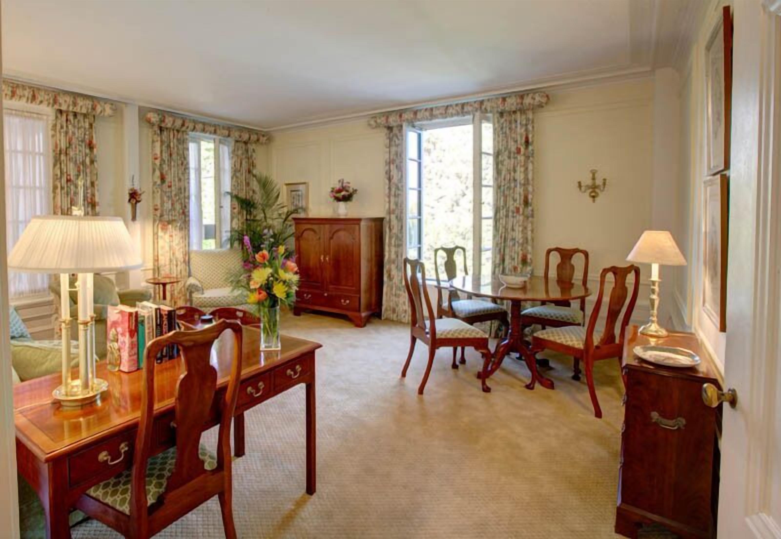 A dining room with a wooden table, chairs, and a cabinet. There are curtains on the windows, and a vase with flowers on the table.