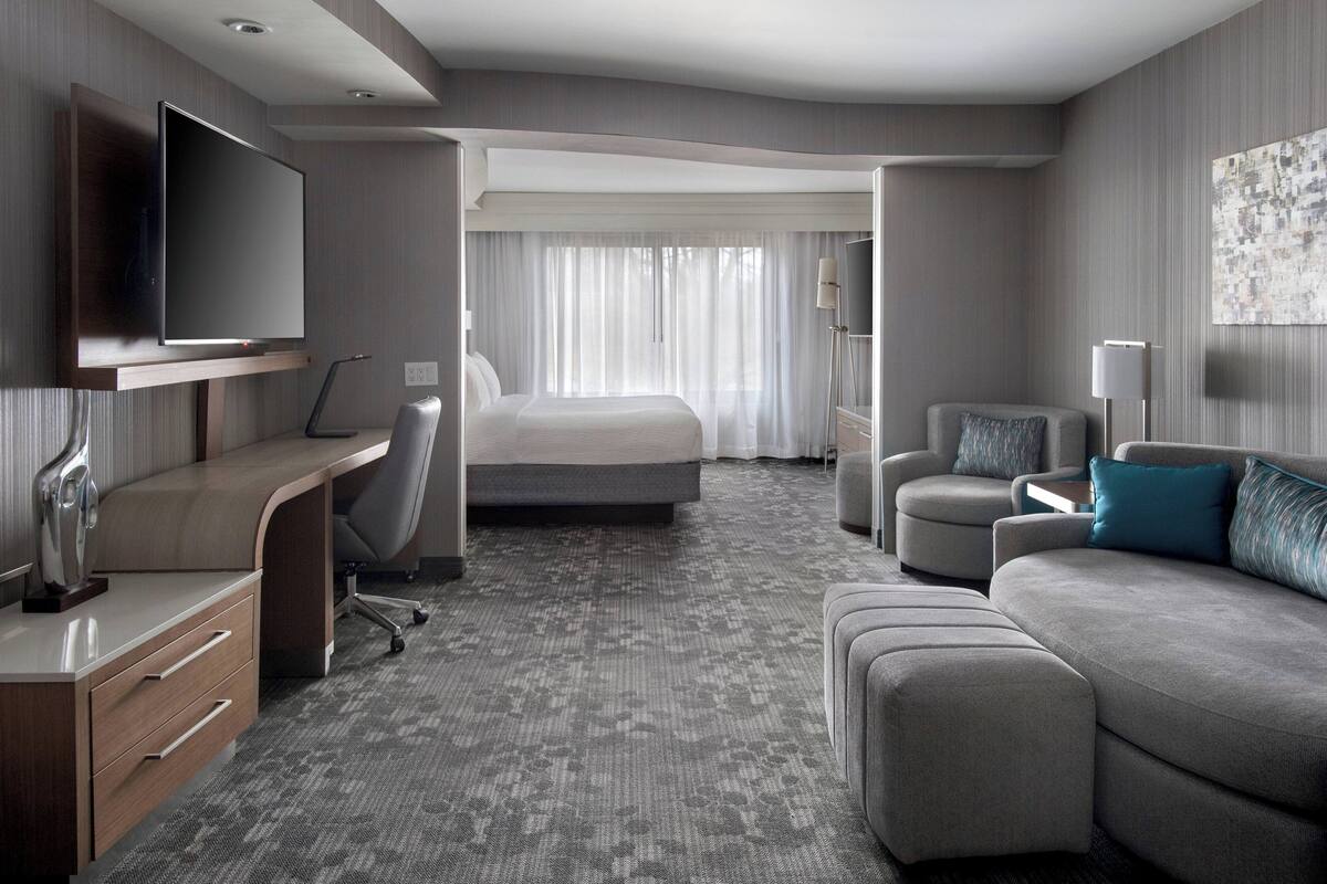 Ein modernes Hotelzimmer mit einem Flachbildfernseher, einem Schreibtisch mit Schubladen, einem bequemen Sofa, einem Bett und einem großen Fenster mit Vorhängen.