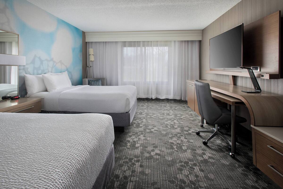 Ein Hotelzimmer mit zwei Betten, einem Schreibtisch, einem Stuhl, einem Fernseher und einem großen Fenster mit durchsichtigen Vorhängen.