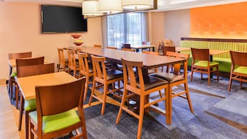 A hotel dining area with multiple wooden tables and chairs, a flat-screen TV mounted on the wall, and a large window with curtains.