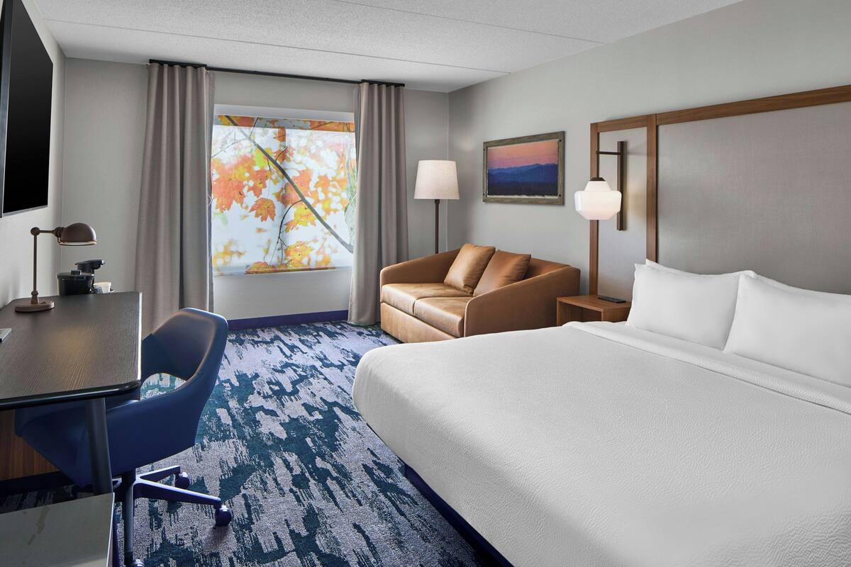 Habitación de hotel con cama, escritorio con silla, lámpara, mesita de noche y ventana con cortinas.