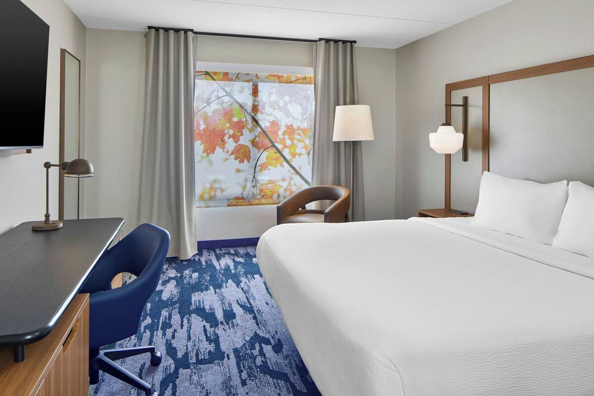Una habitación de hotel con cama, escritorio, silla y un amplio ventanal con cortinas.