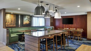 A modern hotel dining area with a green sofa, wooden chairs, a central island, and a flat-screen TV on the wall. - Fairfield Inn by Marriott Rochester East (Webster)