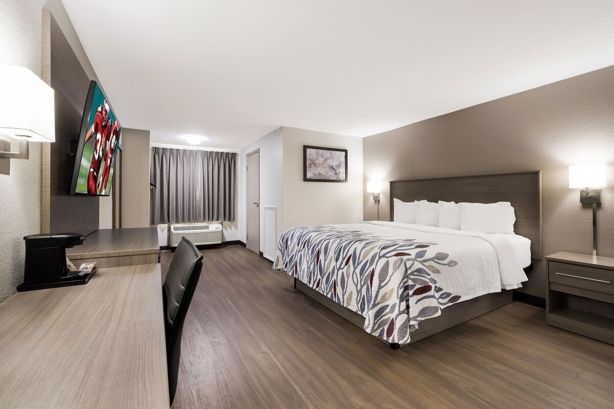 A hotel room with a bed, bedside tables, a desk with a chair, a TV, and a window with curtains.
